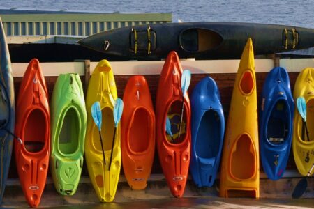 A colorful row of kayaks in various bright colors, including red, orange, yellow, blue, and green, leaning against a wall near a body of water. A black kayak is positioned horizontally above them.