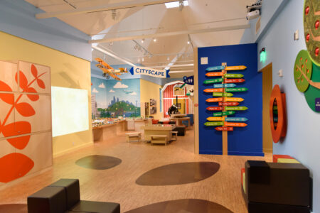 A vibrant children's play area with colorful wall art, a toy cityscape, and a variety of seating. A large blue wall displays colorful, directional signs. A plane model with 