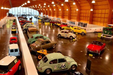 A spacious car museum features a variety of vintage and classic cars on display. The arched wooden ceiling adds warmth, while large windows at the end provide natural light. The cars are arranged neatly in rows on a polished floor.