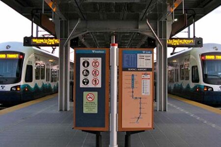 Two light rail trains at a station platform, one on each side. Overhead signs read 