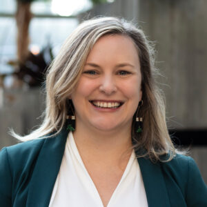 A person with long blonde hair smiling, wearing a green blazer, white blouse, and green earrings stands in a bright space with plants and concrete walls in the background.