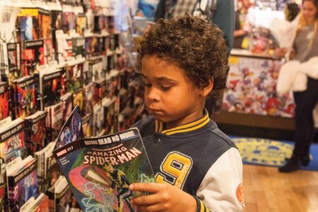 A young child with curly hair browses a Spider-Man comic book in a store filled with colorful comics. In the background, a person in a green jacket and another person are also browsing. The store has wooden floors and bright lighting.