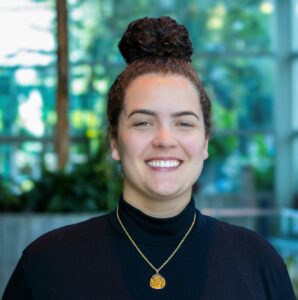 A person with curly hair in a bun, wearing a black turtleneck and a gold necklace, smiles while standing indoors with a blurred background of greenery and glass walls.