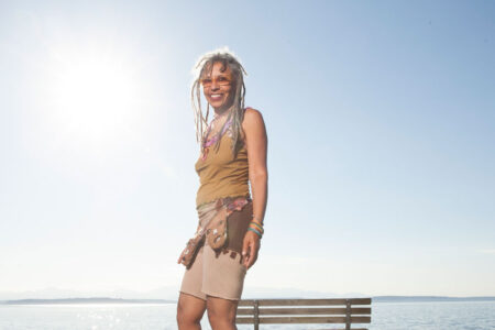 A person with white dreadlocks, wearing sunglasses, a sleeveless top, and a skirt, stands on a bench by a calm sea. The sun is shining brightly in the clear blue sky.