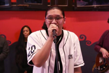 A person wearing glasses and a white baseball jersey is singing into a microphone. Three musicians stand in the background with a red and black wall behind them. One holds a guitar and the others appear to be observing.
