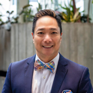 A person wearing a dark suit with a colorful bow tie stands indoors with plants and a concrete wall in the background, smiling at the camera.