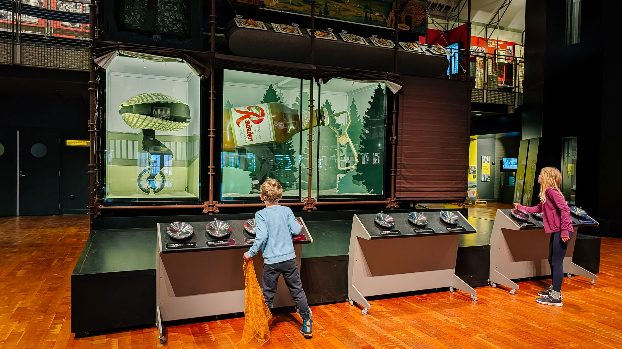 Two children interact with a museum exhibit featuring a large, vintage-style bottle and related graphics. The setting is indoors, with wood flooring and a variety of interactive displays for visitors.