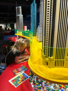 Two young girls eagerly sort through loose lego bricks while examples of professionally-built LEGO towers soar above them at a MOHAI exhibit. 