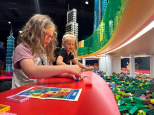 Two young girls construct their own LEGO masterpieces while examples of professionally-built LEGO towers soar above them at a MOHAI exhibit. 