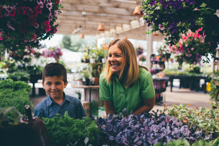A smiling woman and young boy explore a garden center in Woodinville, admiring the myriad of plants. Surrounded by colorful flowers and lush greenery, they soak in the vibrant and cheerful atmosphere.