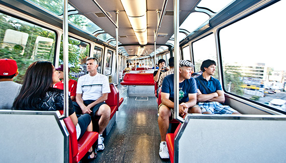 Interior of a train with passengers seated on red seats. People are casually dressed, some engaging in conversation, others looking out the windows. Large windows allow bright daylight to illuminate the cabin, revealing a view of trees and buildings.