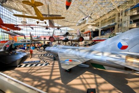 Aviation museum with various aircraft displayed, including a silver jet with a roundel emblem. Several other planes hang from the ceiling. The space is bright with natural light filtering through large windows. Visitors are exploring the exhibits.