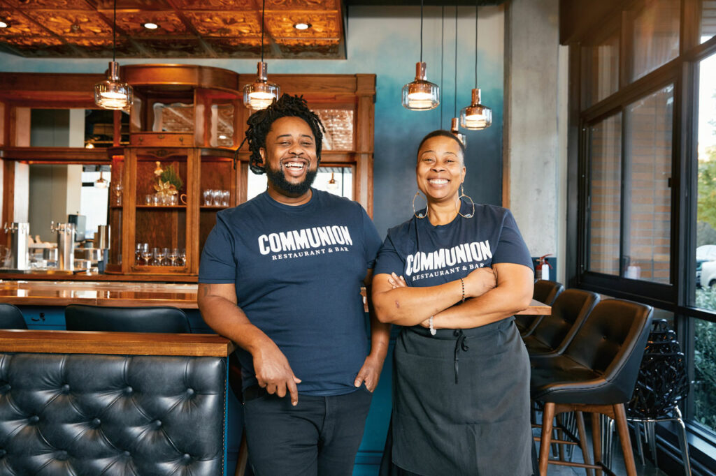 A photo of two people dressed in all black standing in front of a dark wood bar with tufted leather booths.