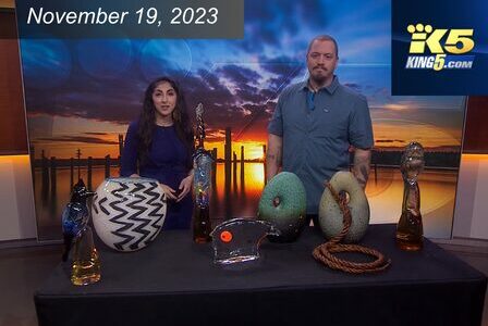 Two people stand behind a table displaying decorative glass sculptures, including vases and abstract shapes. The backdrop features a sunset over a pier. A date and the logo 