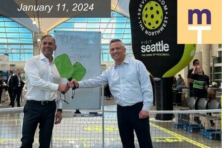 Two men in white shirts holding green pickleball paddles stand by a net in an indoor setting. A sign in the background reads 