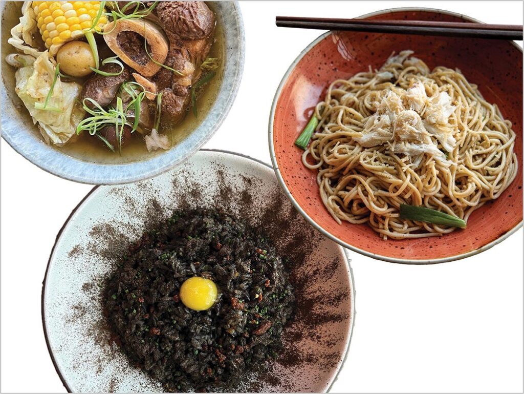 Three dishes are displayed: a bowl of hearty soup with vegetables and corn, a plate of pasta topped with crab meat and garnished with rosemary, and a bowl of black rice with a raw egg yolk in the center. Chopsticks rest on the pasta plate.