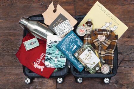 An open suitcase filled with various Seattle-themed items: T-shirts, a flannel shirt, postcards, a cutting board, a metal water bottle, a book, a jar of sauce, coffee packaging, and a yellow poster. The suitcase is placed on a wooden floor.