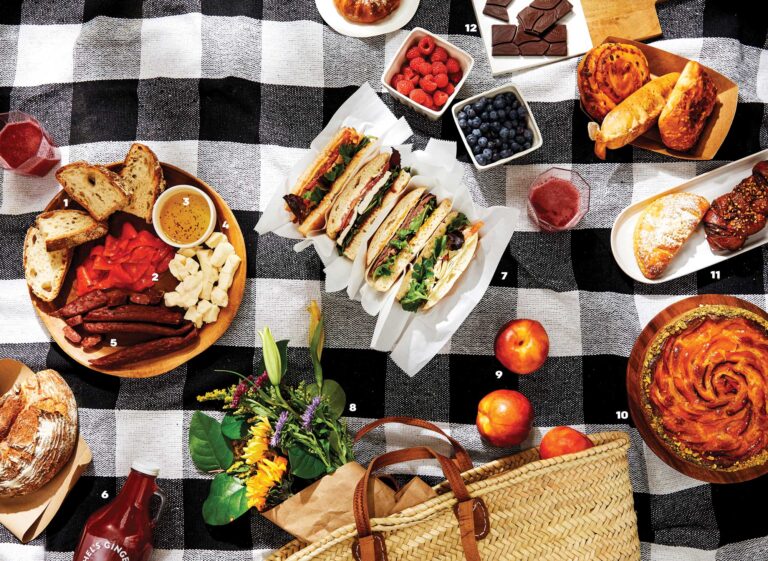 A picnic spread on a checkered blanket featuring sandwiches, fruit, chocolate, pastries, sausages, bread, cheese, and a bouquet of flowers. A wicker basket and a bottle with a red label are also visible.
