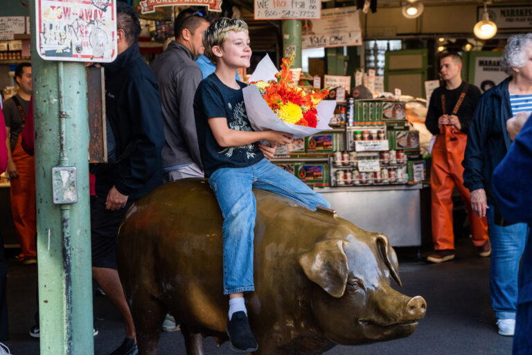 A young boy holding a bouquet of flowers sits on a bronze pig statue at an indoor market. People and market stalls are visible in the background.