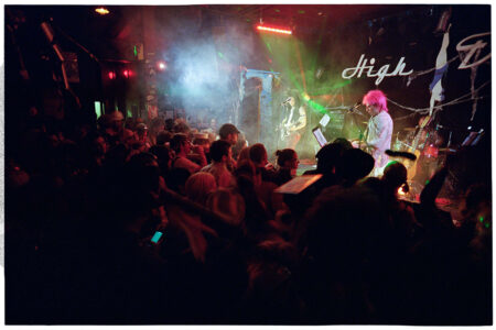 A band performing on stage in a dimly lit venue, with colorful lights and smoke effects. The audience is gathered closely, enjoying the music. The band's name, partially visible, is displayed in the background.