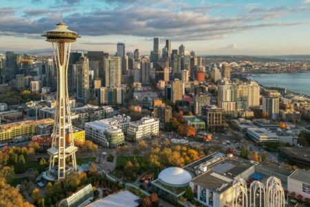 Aerial view of Seattle's skyline with the Space Needle in the foreground. The cityscape features a mix of skyscrapers and buildings, set against a backdrop of a partly cloudy sky and distant mountains. Autumn trees add color to the scene.