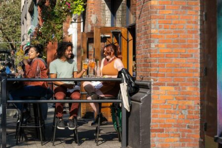 Three people sit outside a brick-walled café, enjoying drinks and lively conversation. Two hold glasses of beer, while the third sips a yellow beverage. The ambience is warm with hanging flowers and sunlight.