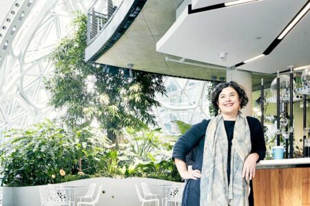 A person stands smiling beside a counter in a modern, airy indoor space with large glass windows and lush green plants. The setting appears to be a cafe or lounge area with seating and natural light.