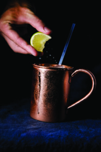 A hand squeezes a lime wedge over a copper mug with ice and a straw, capturing droplets in motion. The mug, slightly frosted, stands out against a dark background.