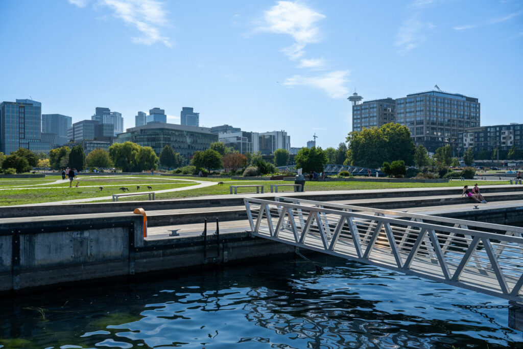 Scenic bridge crossing the calm shores of Lake Union, with a green lawn, trees, and blue sky in the background. 