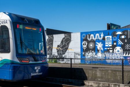 A Sound Transit light rail train approaches a station, with vibrant street art decorating a nearby wall. The mural features abstract patterns and various symbols in black and white, contrasting against a bright blue background under a clear sky.