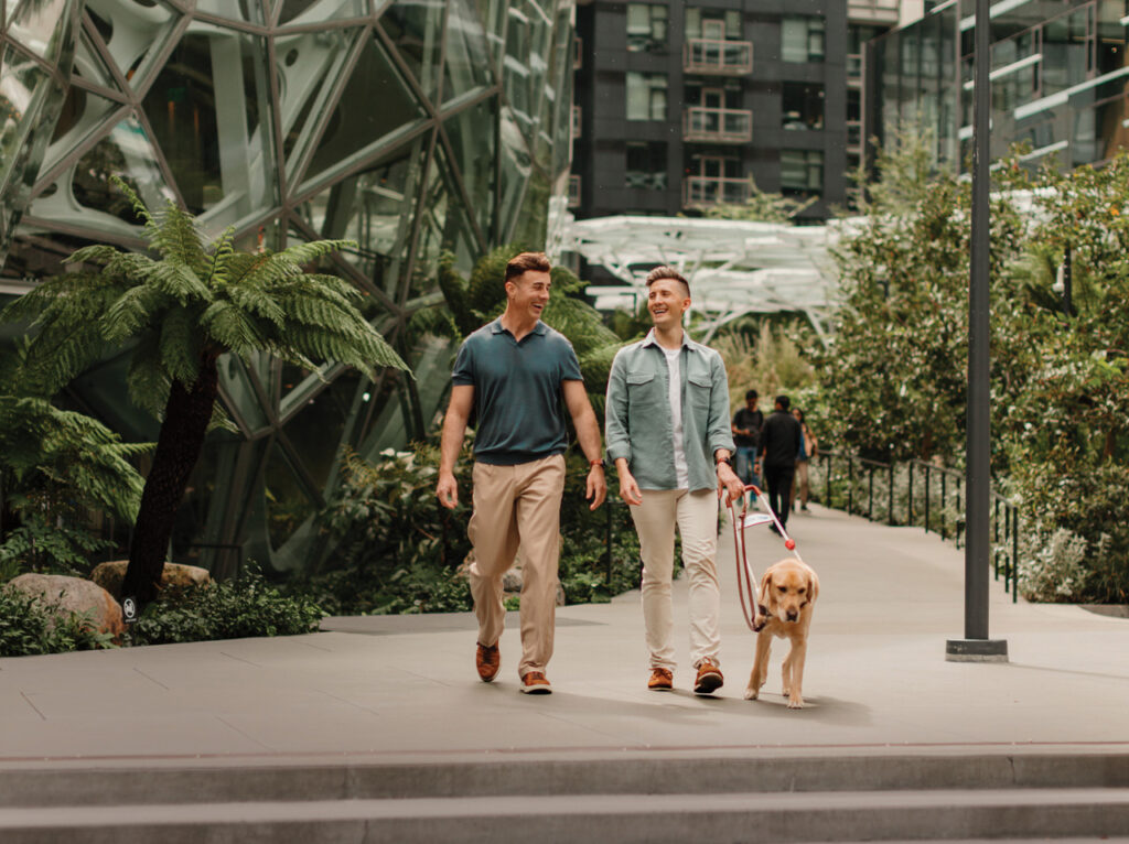 A photo of two people walking next to the Seattle Spheres. The person on the left has a short sleeve green polo and khaki pants on. The person on the right has a green long sleeve button up with a white t-shirt and khaki pants on. The person on the right holds the harness for a yellow colored guide dog.