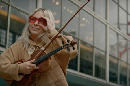 A person with long blonde hair wearing red glasses smiles while holding a violin outdoors. They are standing in front of a large building with reflective windows.