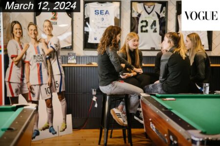 A group of people sitting on stools and chatting in a sports-themed room with framed jerseys and pool tables. The date 