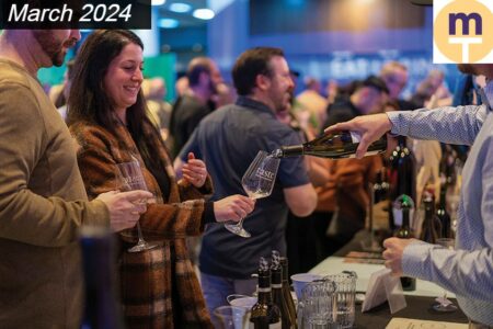 People gather at a wine-tasting event. A person pours wine into a glass held by a smiling attendee. Others engage in conversation, with bottles displayed on a table. A 