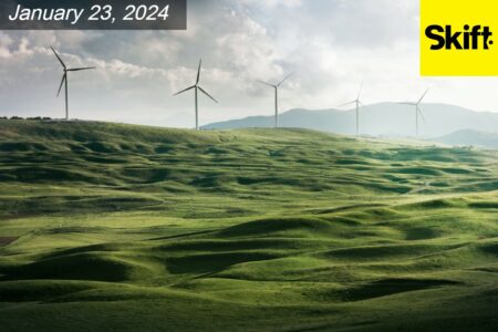 A scenic view of green rolling hills with five wind turbines perched on them, set against a cloudy sky. The date 