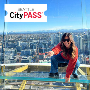A person in a red sweater and sunglasses crouches and points on a glass platform high above a cityscape. The Seattle CityPASS logo is in the top left corner. Skyscrapers and mountains are visible in the background.