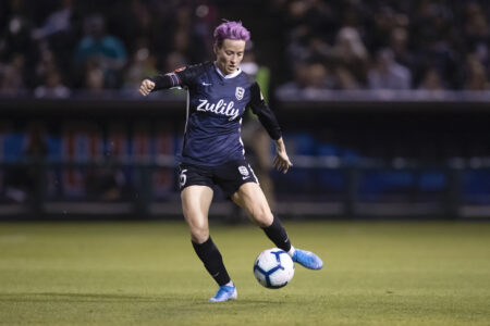 A soccer player with short purple hair, wearing a dark jersey and shorts, is in motion on the field during a game. She is in the midst of kicking a white and blue soccer ball. The background shows a blurred audience.