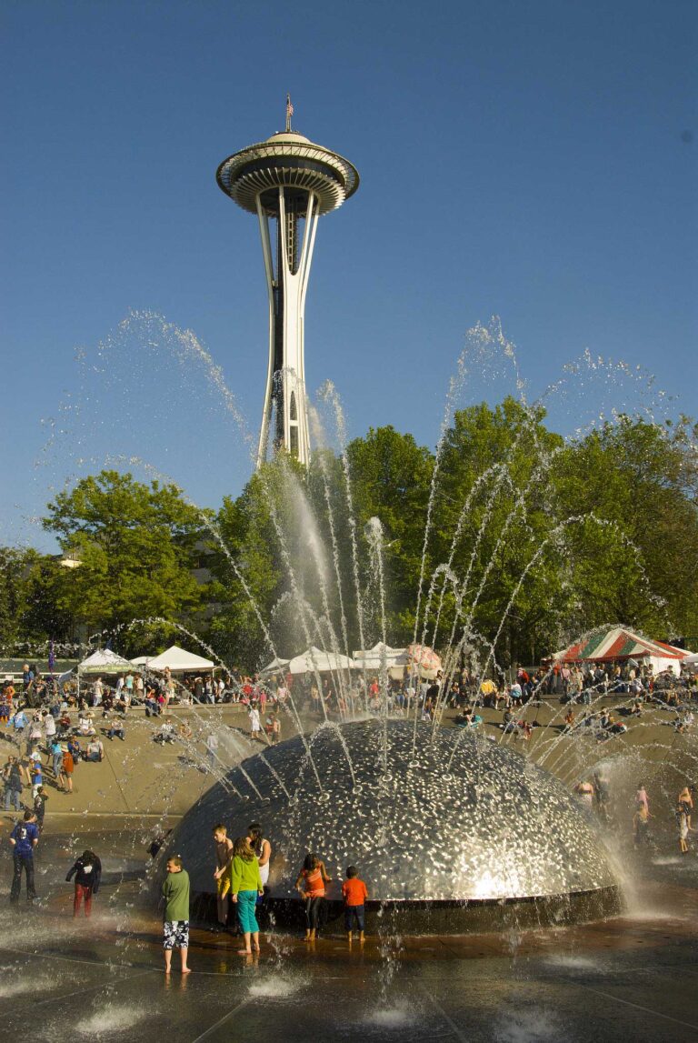 Children play in a large, dome-shaped water fountain on a sunny day. The Space Needle towers in the background. People relax on the grass while tents are set up nearby, suggesting a fair or event. The sky is clear and blue.
