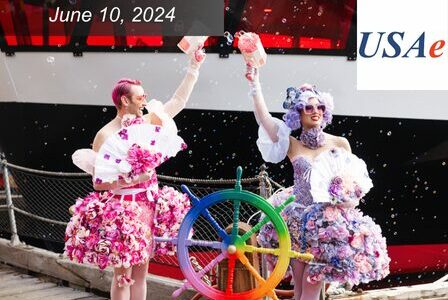 Two drag queens wearing pink and purple flower dresses blow bubbles by a rainbow ship wheel.