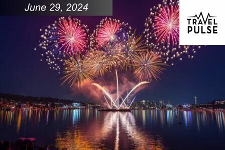 Purple and red fireworks explode over a body of water, which has a colorful reflection.