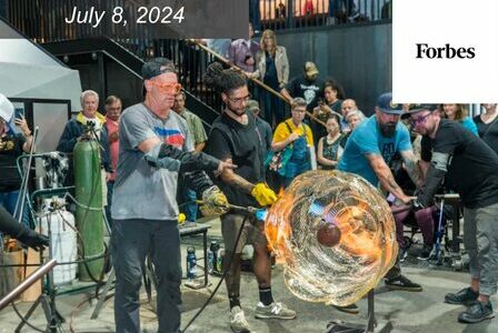 A white man and a Black man showcase a glass blowing demonstration to a crowd.