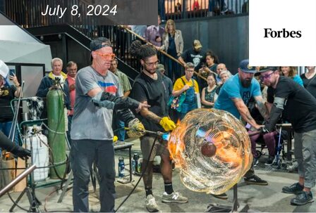 A white man and a Black man showcase a glass blowing demonstration to a crowd.