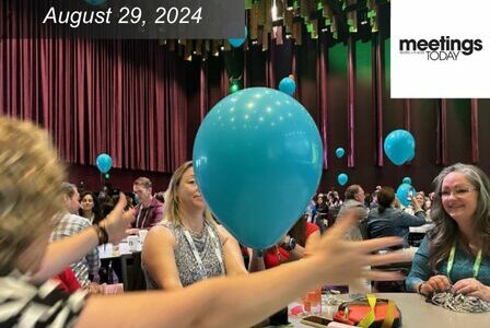 A lively conference scene with people seated at tables, engaging and smiling. A large blue balloon is prominent in the foreground. The room is brightly lit with decorative elements. A caption reads 