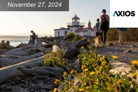 A group of people and a dog walk along a driftwood-strewn path by the sea, with a lighthouse in the background. Yellow flowers dot the foreground.