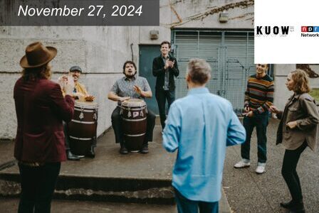A group of people stand outside in front of a concrete wall, with two people playing drums. Others are clapping and singing.