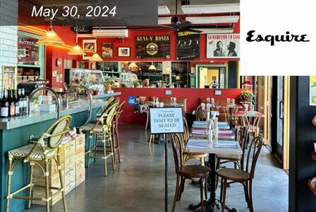 A vintage-themed restaurant interior with eclectic decor, colorful posters, and a mix of wooden and woven chairs. A sign on a table reads 