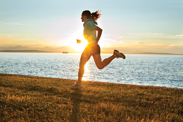 A person is running along a grassy shoreline with the sun setting in the background. The silhouette is illuminated by the sun, creating a warm glow. A body of water and distant mountains are visible under the colorful sky.