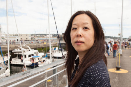 A woman stands on a marina dock, surrounded by moored boats and calm water. She gazes into the distance, with a partly cloudy sky overhead and scattered people walking in the background.