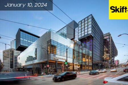 City street view showing a modern glass and steel building with traffic in the foreground. The date 
