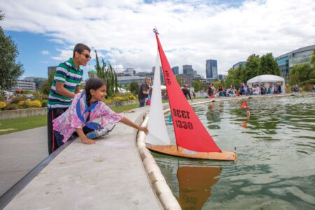 A young girl in a colorful outfit reaches for a model sailboat in a pond, aided by a man in a striped shirt. The scene unfolds against the backdrop of the vibrant South Lake Union neighborhood, where city buildings tower and people bask under the sunny park skies.